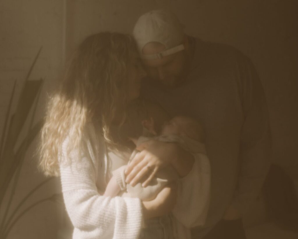 First-time mother and father embrace while cradling their newborn baby in morning light