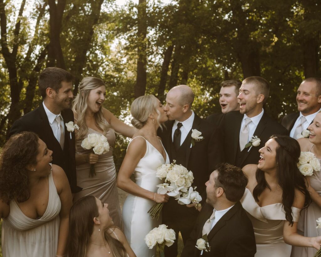 The bride and groom kissing while surrounded by their cheering bridal party members. 