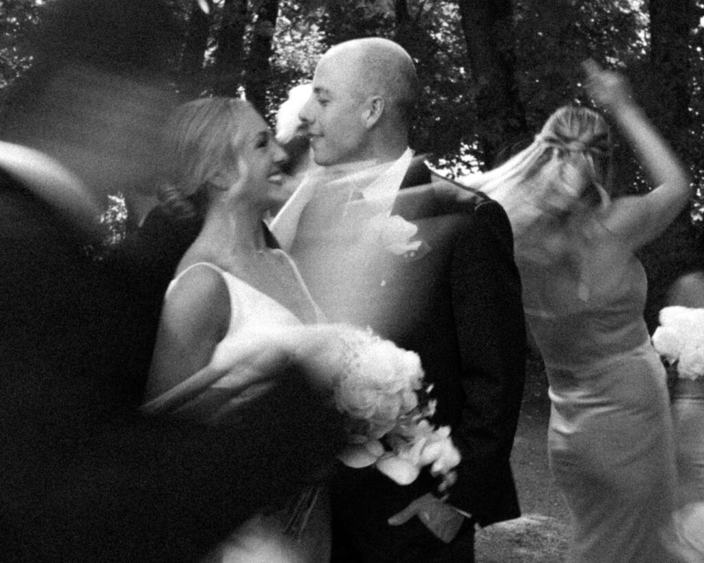 A slow shutter black and white image of the bride and groom smiling at each other while their bridal party members dance around them.  