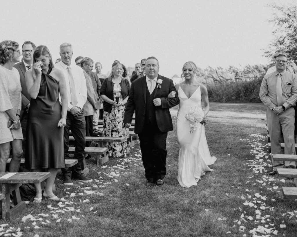 Black and white photograph of the bride and her dad make their way down the aisle while guests watch.