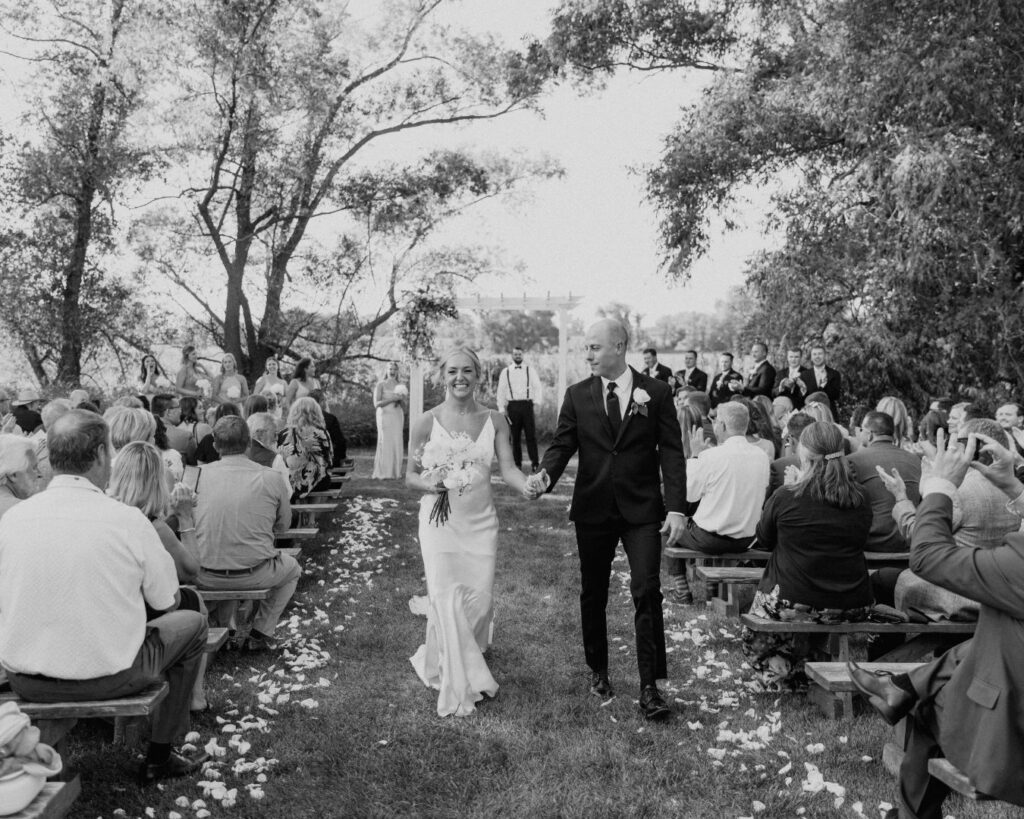 A black and white image of the bride and groom processing down the aisle while their family and friends cheer after being announced as husband and wife for the first time.