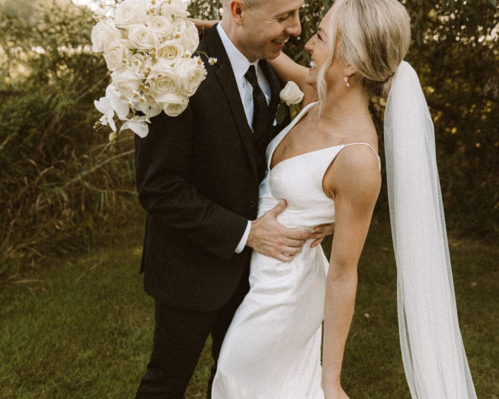 An up close image of the bride and groom wrapped in each others arms with her bouquet of white roses and orchids and smiling at each other.