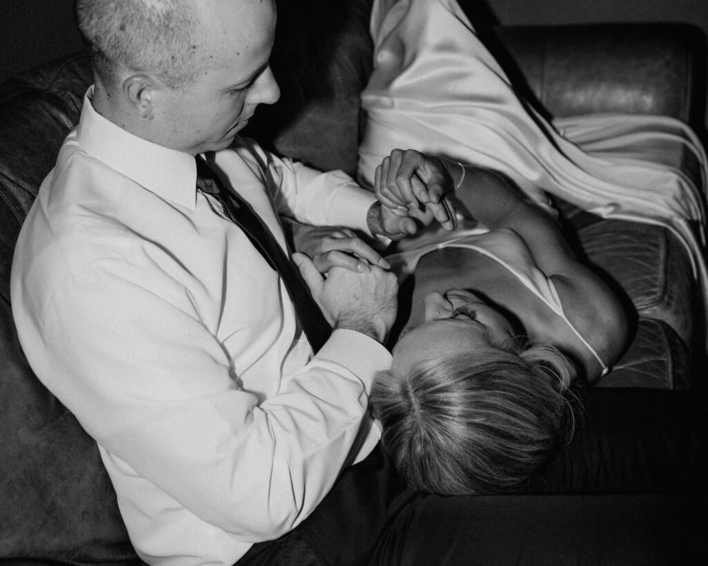 A black and white image of the bride and groom snuggled up on the couch in the grooms suite, playing with each others hands.