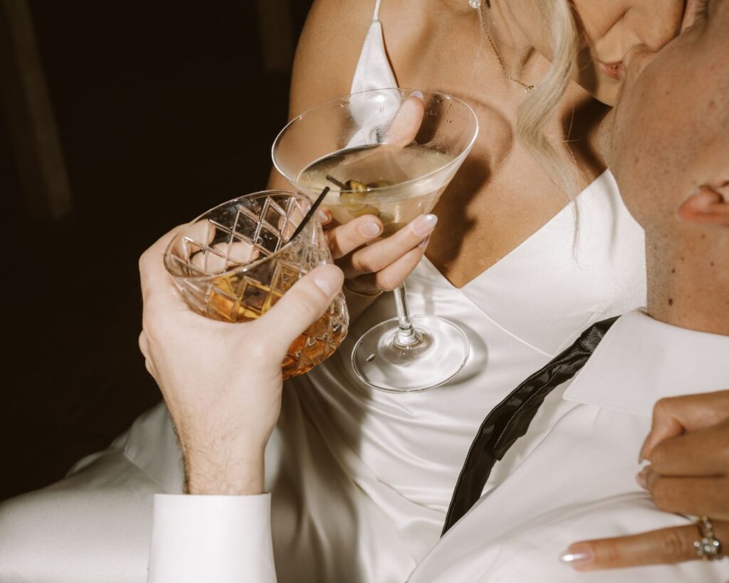 The bride sitting on the groom's lap and kissing him while holding on their favorite drinks- his an old fashioned and hers a martini. 