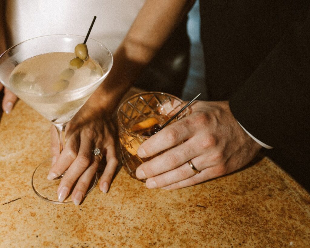 Photo of the bride and groom's ring hands holding their favorite drinks on a table in the groom's suite. His an old fashioned, hers a martini. 