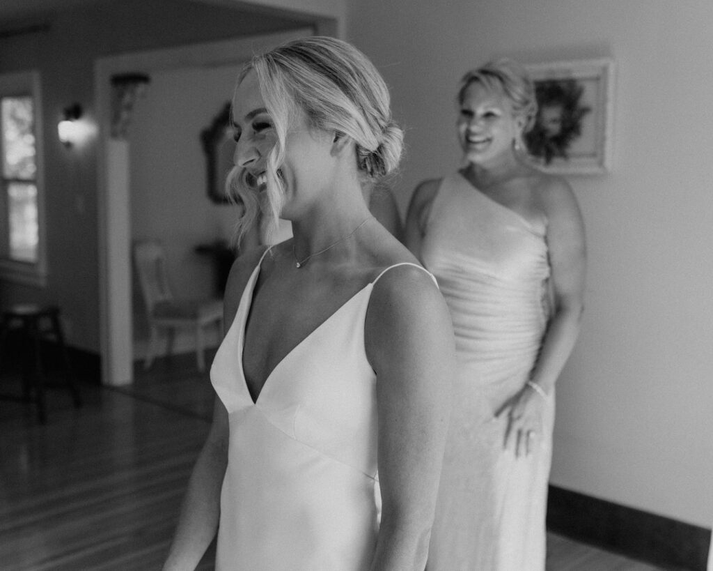 Black and white image of the bride smiling in the mirror after putting on her dress while her mom and sister smile as they watch from behind.