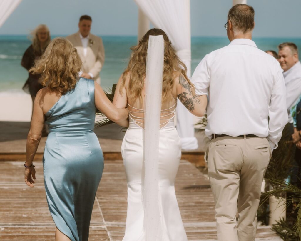 An image from the back of the bride being walked down the aisle by her parents toward her groom during their beach wedding in Costa Mujeres, Mexico.