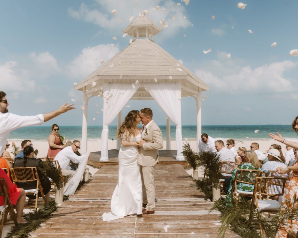 The bride and groom kissing in the middle of the aisle as guests throw white rose petals shortly after they were announced as Mr. and Mrs. with the ocean in the background at their Costa Mujeres, Mexico wedding.