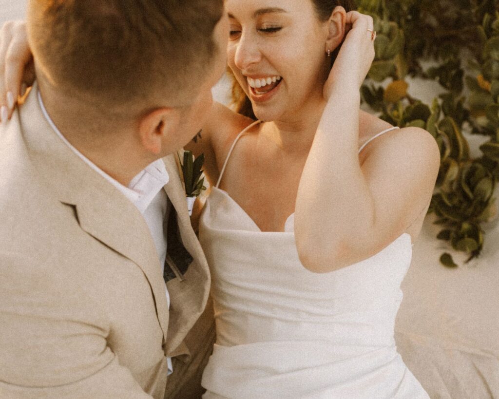 Up close image of the bride sitting in the grooms lap in the sand and giggling. 