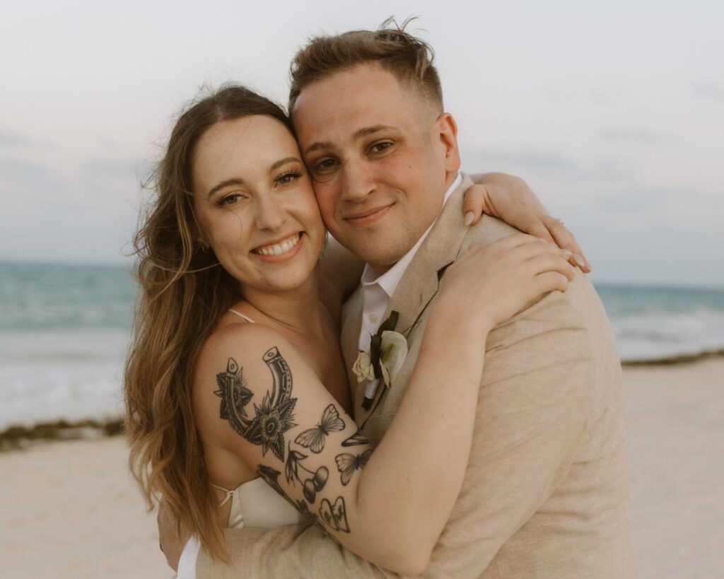 The bride and groom smiling at the camera on their wedding day. The ocean waving behind them. 