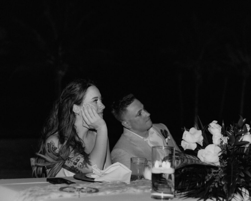 Black and white image of the bride and groom sitting at their table listening to speeches. The bride is wearing a teary expression an the groom a soft smile. 