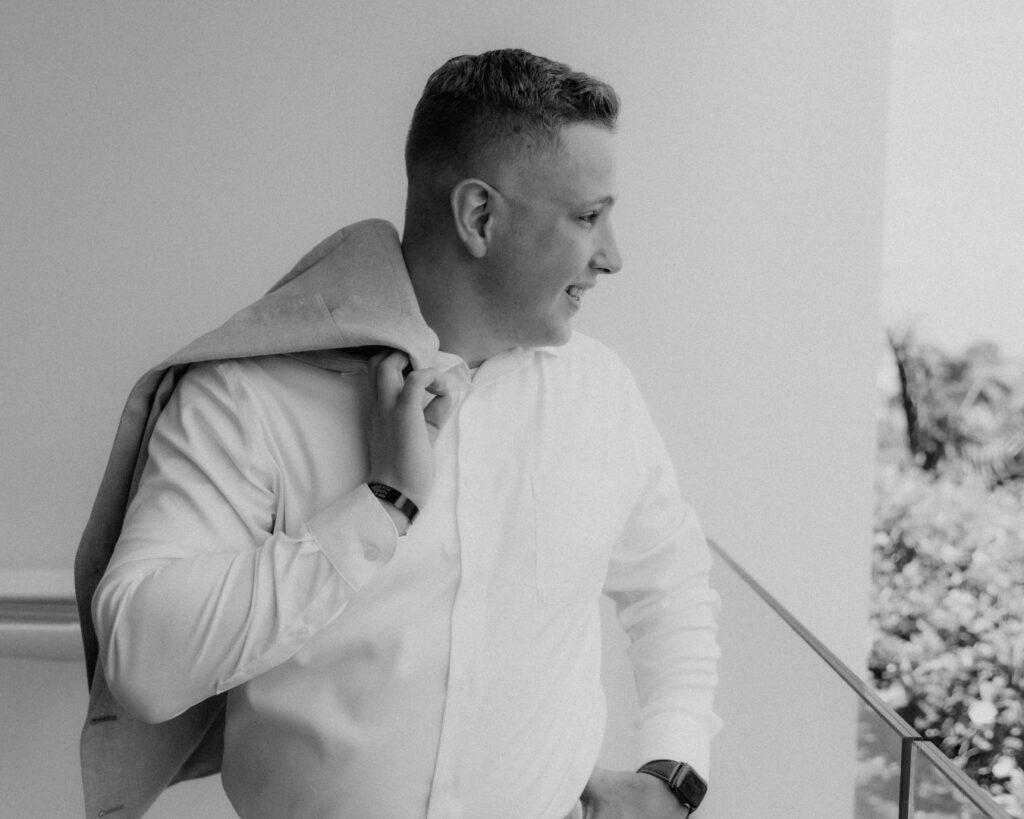 A black and white image of the groom  smiling out over the patio with his jacket slung over his shoulder. 