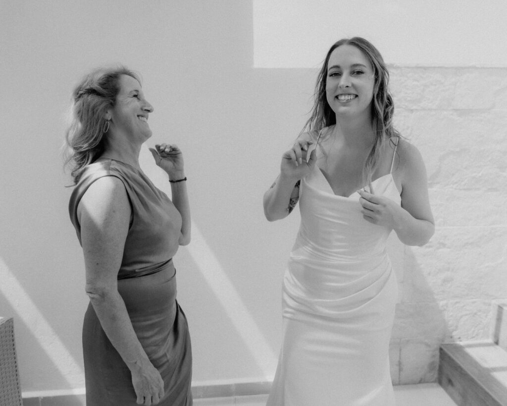 Black and white image of the bride and her mom laughing after putting on her wedding dress. 