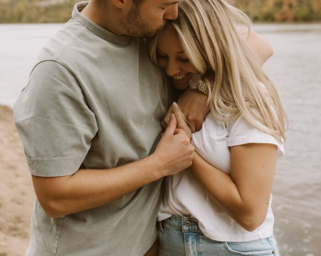 Engaged couple snuggles in tight to one another in front of a river while he kisses her on the temple