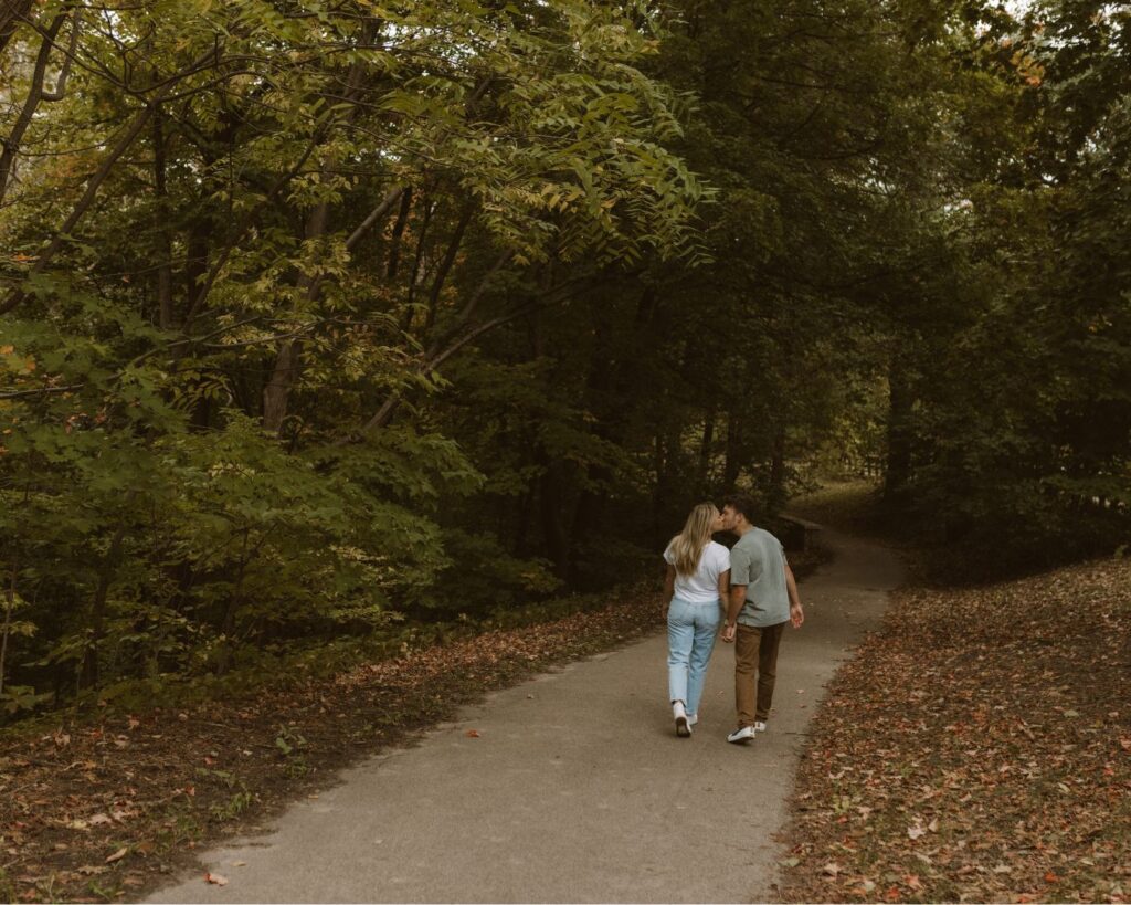 Couple walking down a sidewalk and sneaking a quick kiss while surrounded by green trees during their engagement session. 