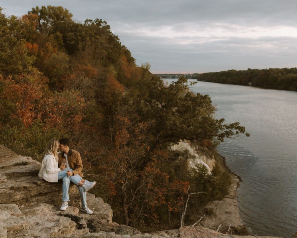 Wide shot of engaged couple sitting  snuggled up on a bluff overlooking the river and fall tree while sipping champagne from their glasses. 