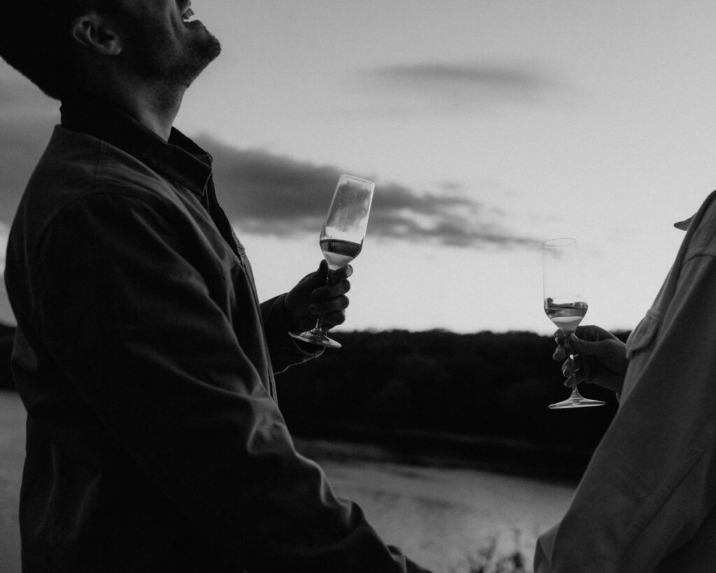 Up close black and white image of couple holding hands and holding their champagne glasses while laughing during their engagement session. 