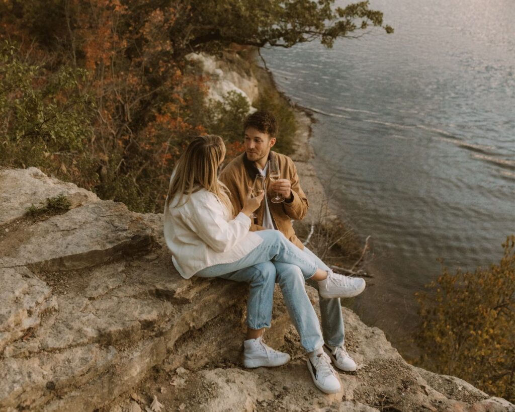 Up close shot of engaged couple sitting  snuggled up on a bluff  and clinking their glasses of champagne together while looking at one another. 