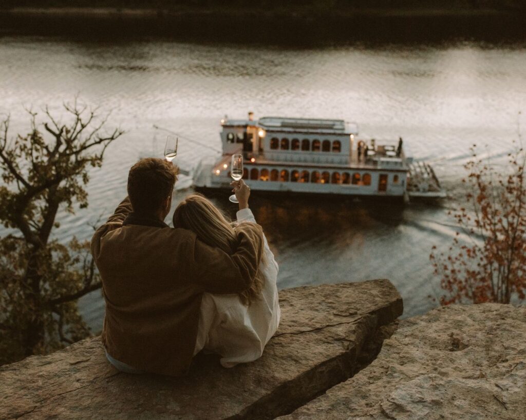 Couple sitting on the edge of a bluff overlooking the river and both putting their glasses in the air to toast to the adorable river boat passing below during their engagement session. 