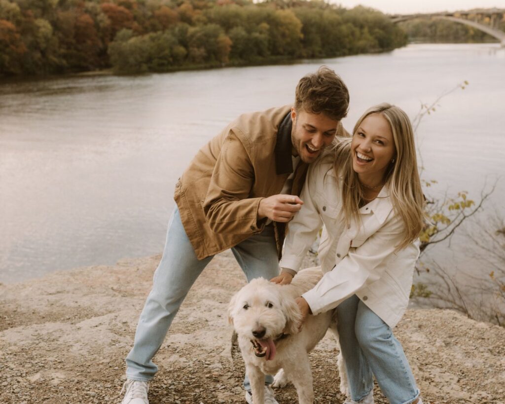 Engaged couple standing on a bluff overlooking a river and laughing as a stranger's labradoodle makes himself comfortable by squeezing right in between them. 