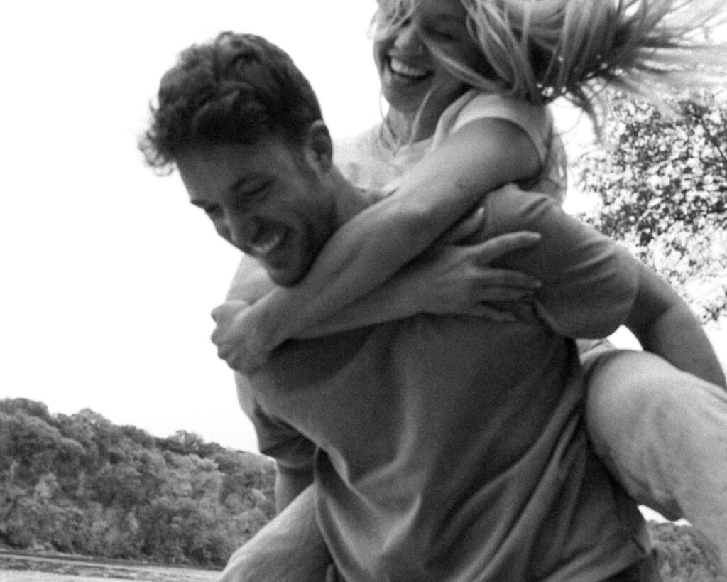 Blurry black and white image of newly engaged couple. He is giving her a piggy back and tossing her around so her hair flies while they both smile. 