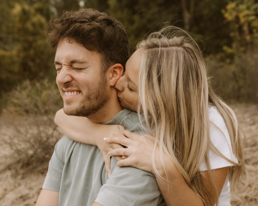 Couple sits on the beach, her behind him and he squints his eyes and smiles as she kisses him on the back of the neck. 