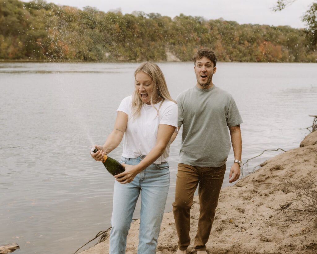 Couple pops champagne with surprised faces while standing on a beach overlooking a river during their engagement session.