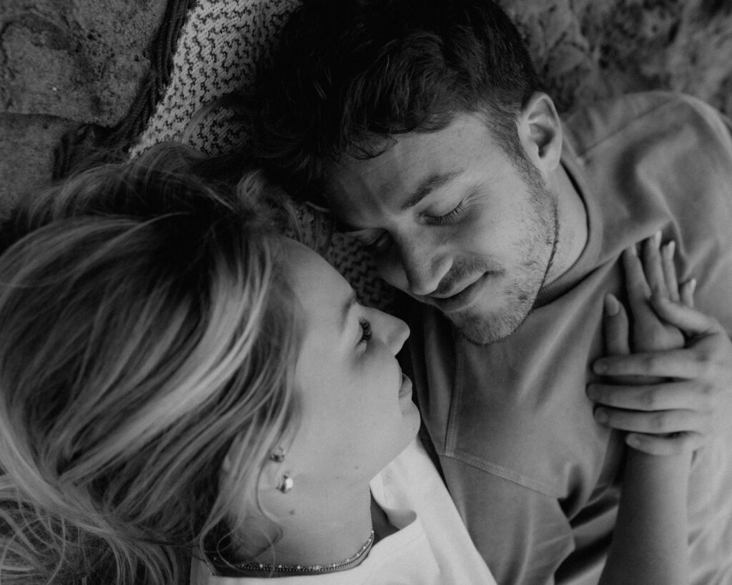 Black and white photo of an engaged couple laying on a blanket on the beach just before kissing. 