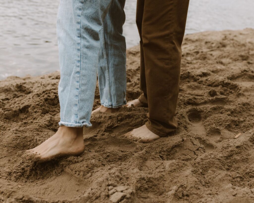 Image of the engaged couples feet covered in sand as he hugs her from behind. 