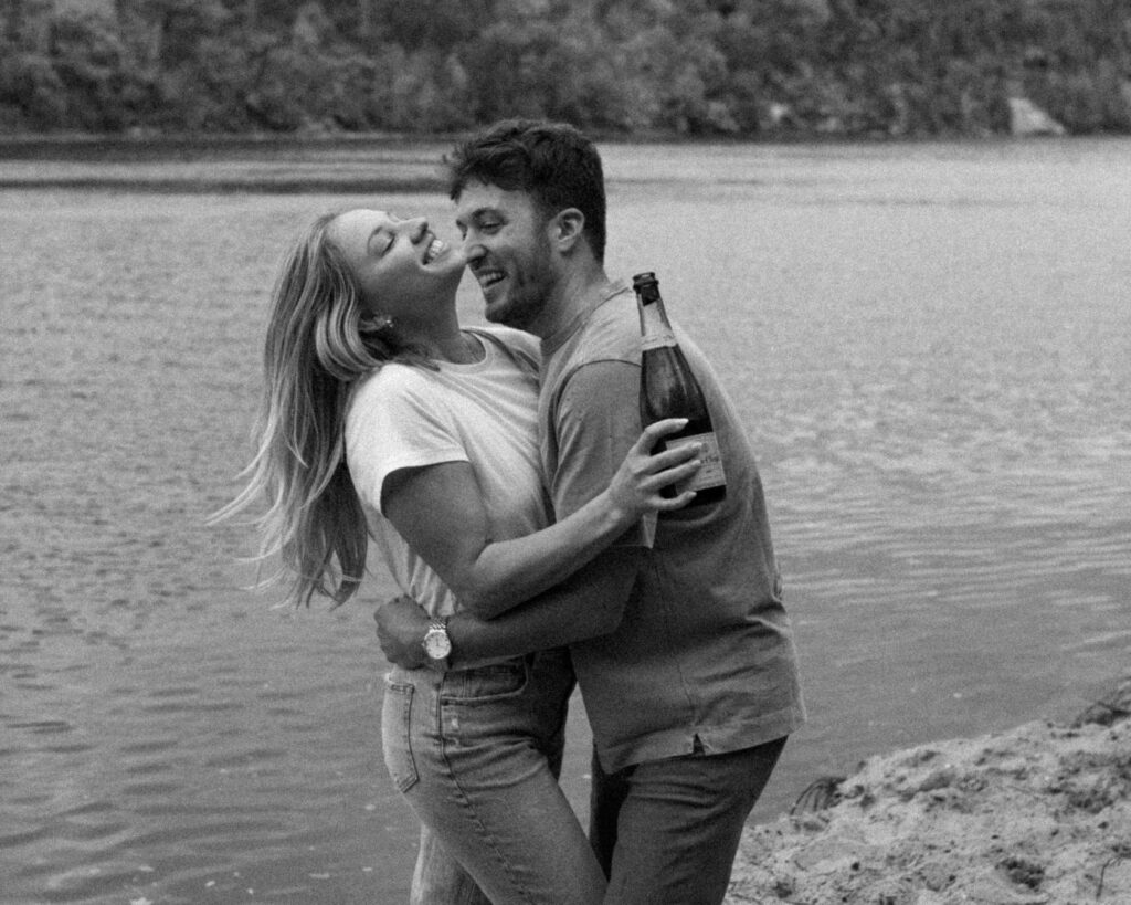 Black and white image of engaged couple on the beach hugging each other and smiling while she holds a bottle of champagne. 