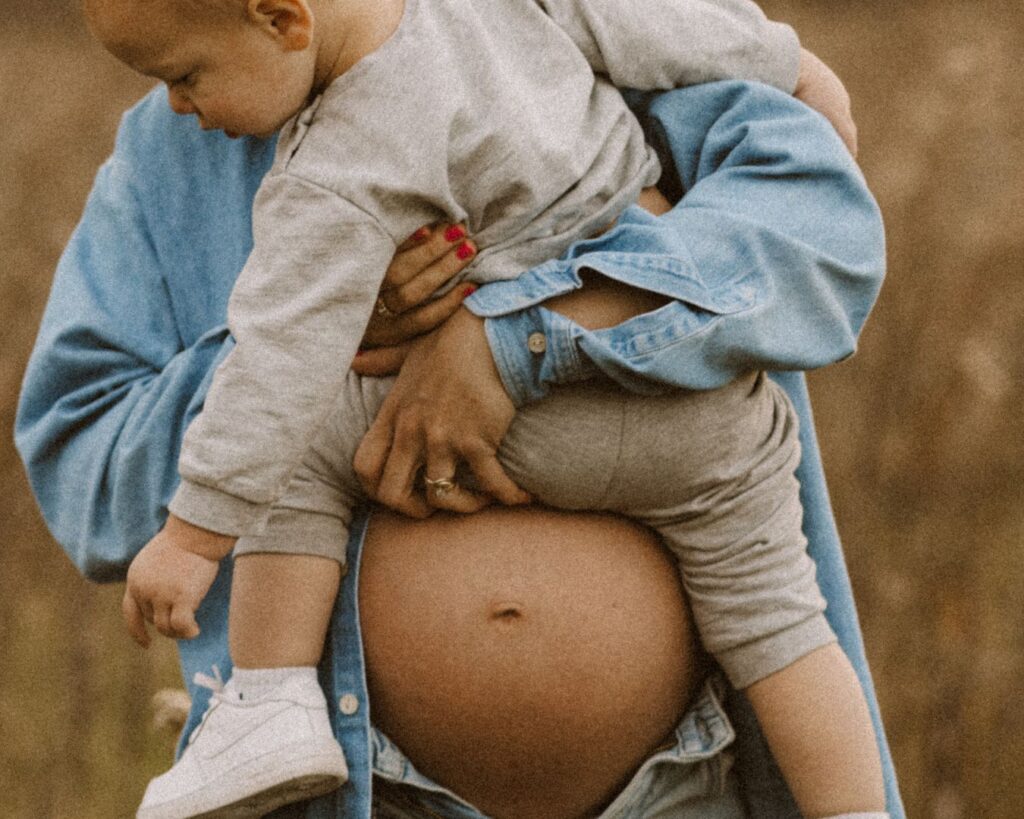 Pregnant mom shows off her bare belly by wearing an open button down denim shirt while holding her toddler. 