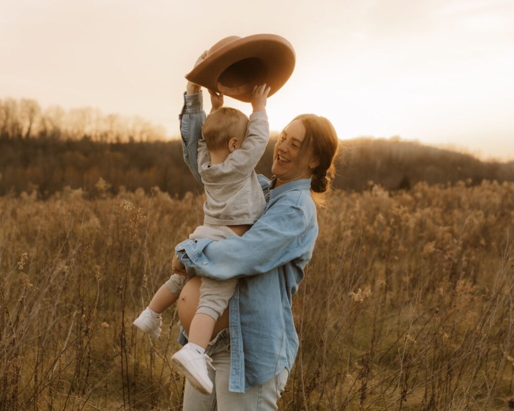 Pregnant mom holding her toddler while he attempts to put her pink wide brimmed hat on to her head at Minnesota golden hour.