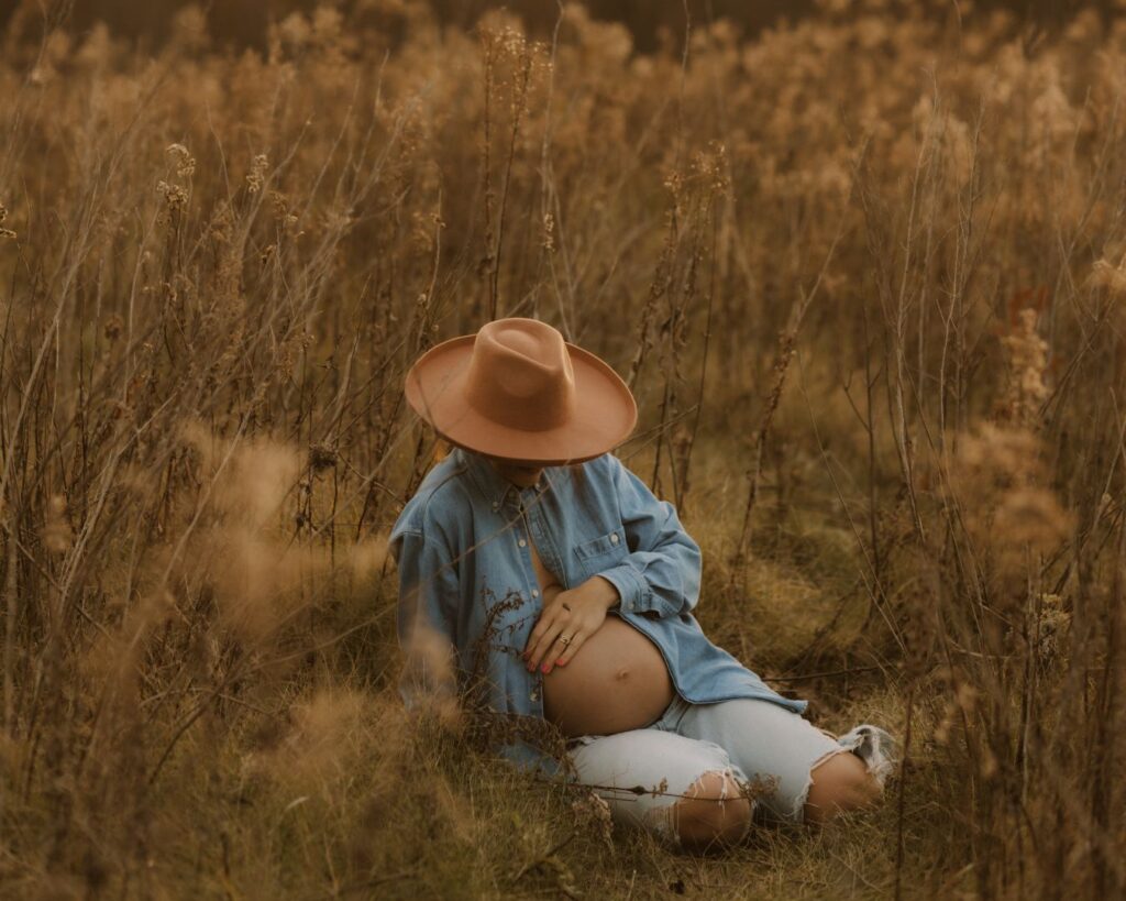 Pregnant mom during her maternity session, sitting on the ground and caressing her belly. She looks down at her belly while she does this, the wide brimmed hat she's wearing hiding most of her face. 