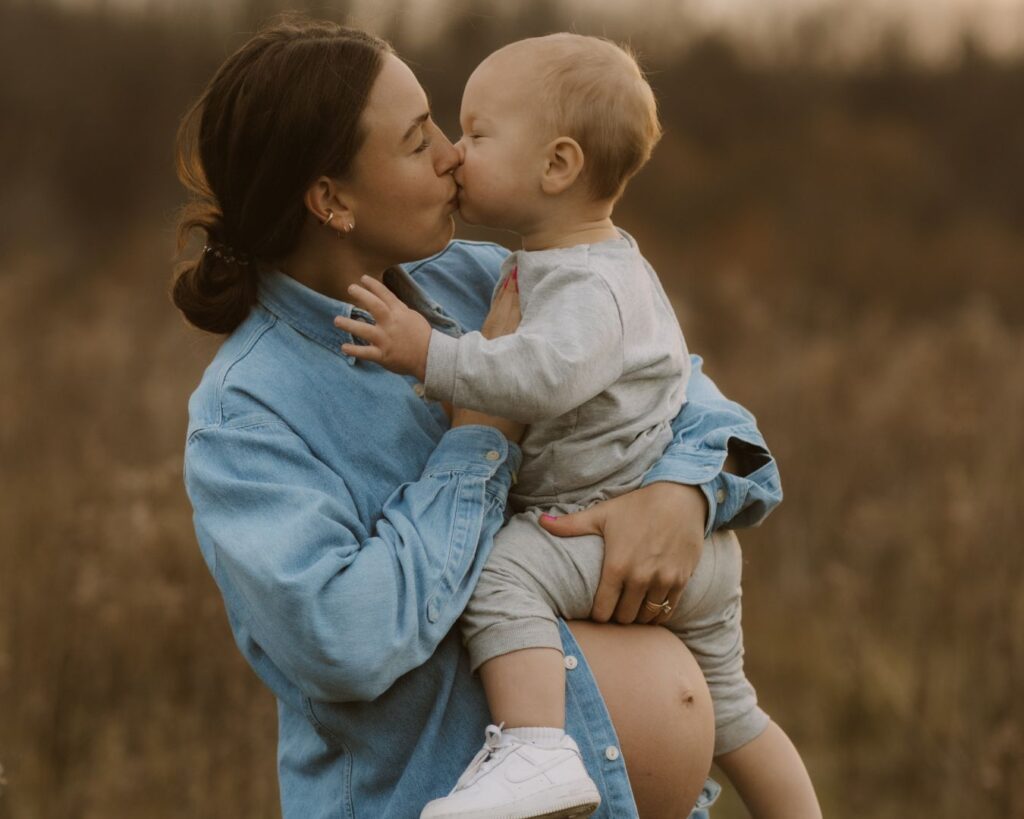 Pregnant mom shows off her bare belly by wearing an open button down denim shirt and giving her toddler a kiss while holding him.