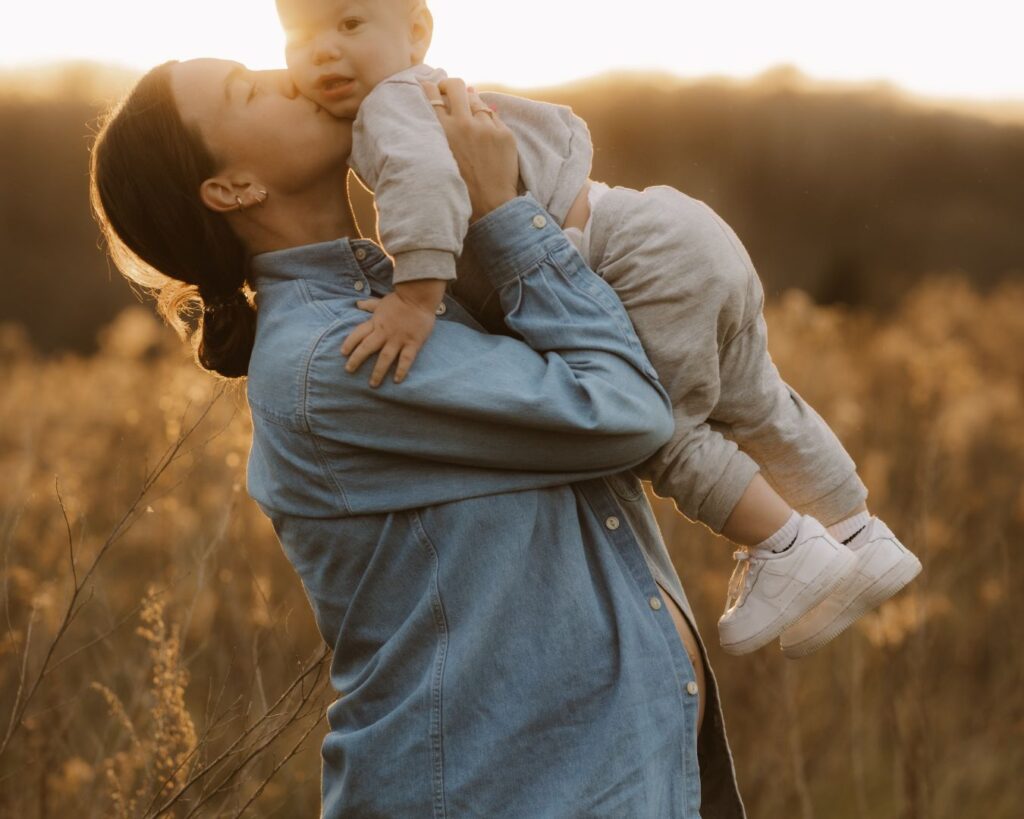 Pregnant mom holds her toddler up, kissing his cheek as they are surrounded by the Minnesota golden hour light of the setting sun. 