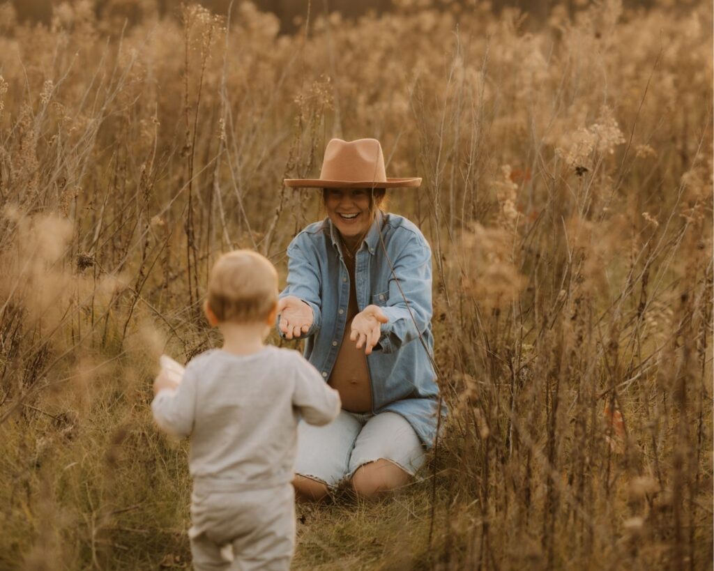 Pregnant mom kneels on the ground with open arms welcoming her toddler as he runs towards her. 