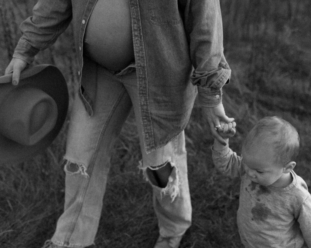 Up close black and white image of pregnant mom holding her messy toddler's hand while they walk through a field at Minnesota golden hour.