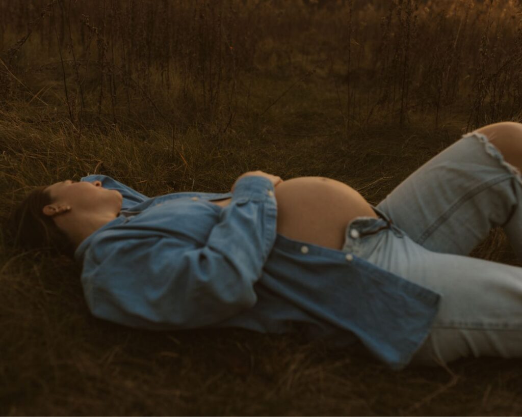 Pregnant mom laying in an open field as the sun goes down and softly lights up her bare belly. 