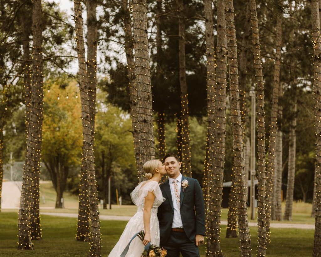 Bride kissing the groom on the cheek in front of the tall pine trees that are now lit up with warm soft twinkle lights. 