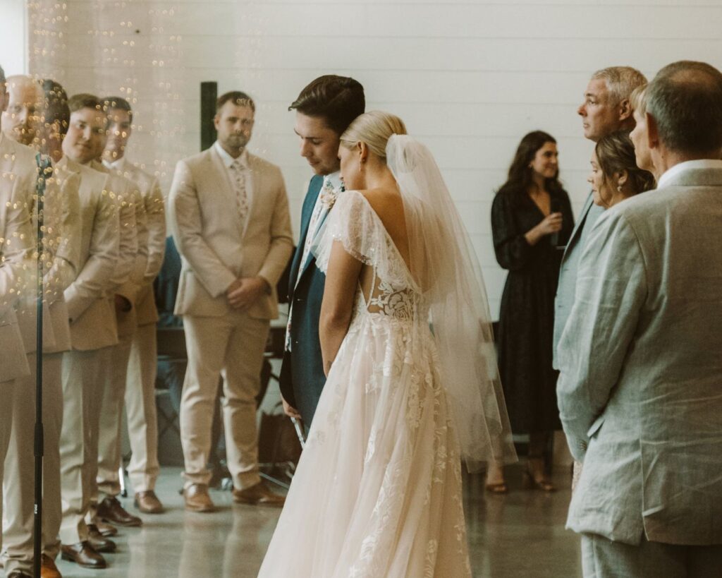 Image of the bride and groom durning their ceremony through a window. She has her head laid on his shoulder and they are surrounded by their bridal party and guests. 