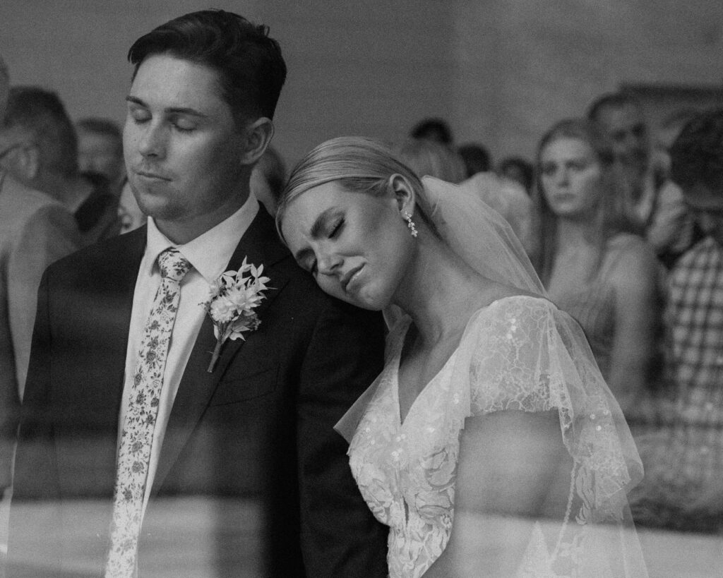 Black and white image of the bride and groom through the window with the bride's head on the groom's shoulder during their wedding ceremony. 