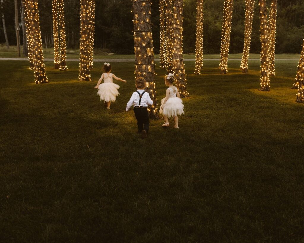 Tow flower girls and one ring bearer running through the twinkle light pine trees as it get's dark. 