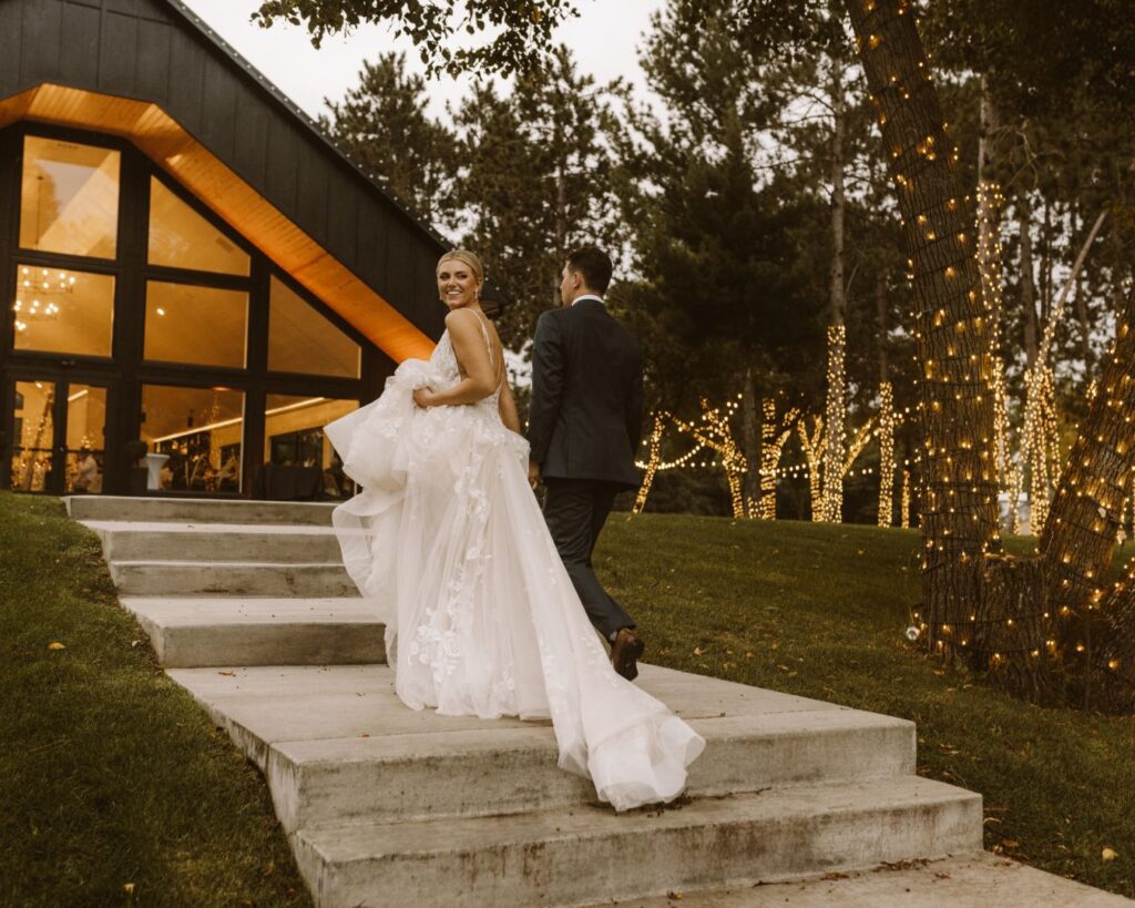 The bride and groom making their way back into their warmly lit venue with the twinkle lights behind them. The bride is looking back over her shoulder towards the camera as they walk hand in hand.