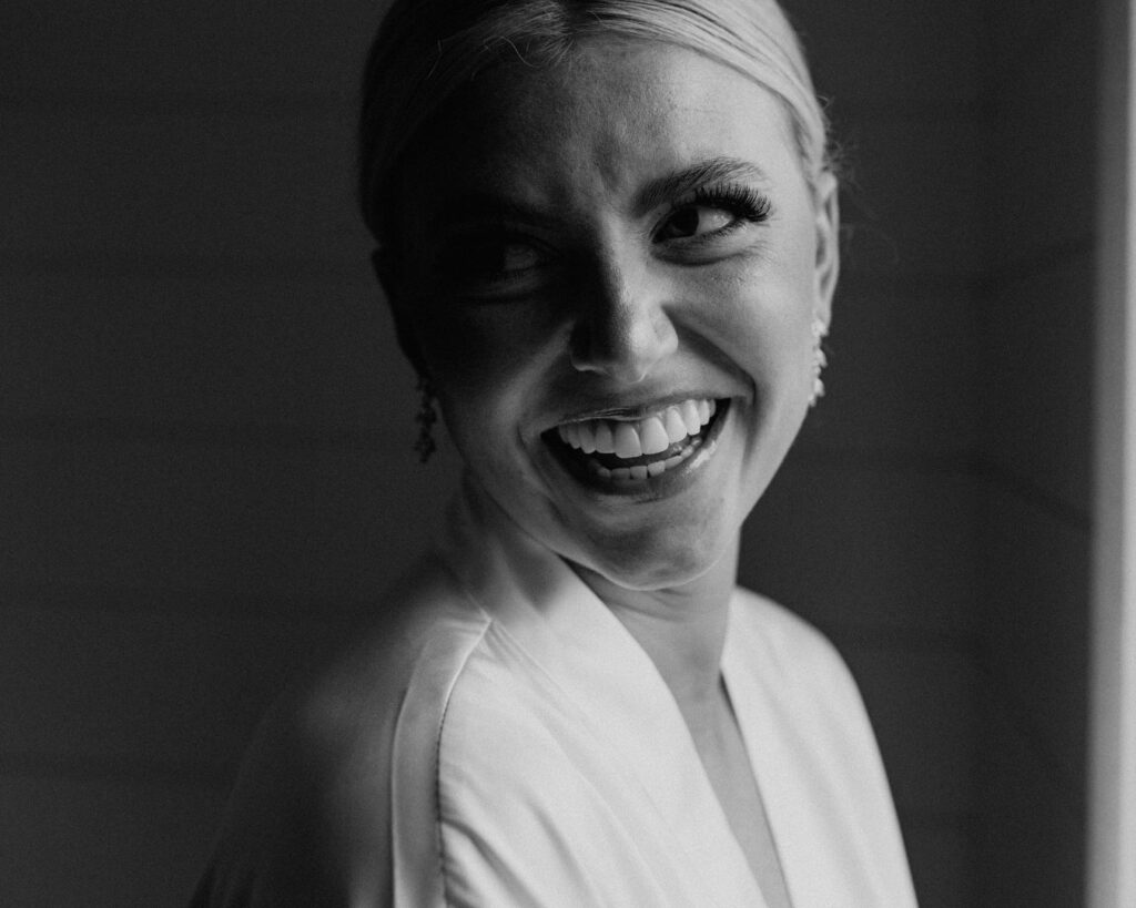 Black and white image of the bride smiling over her shoulder in the light of a window before putting on her wedding dress 