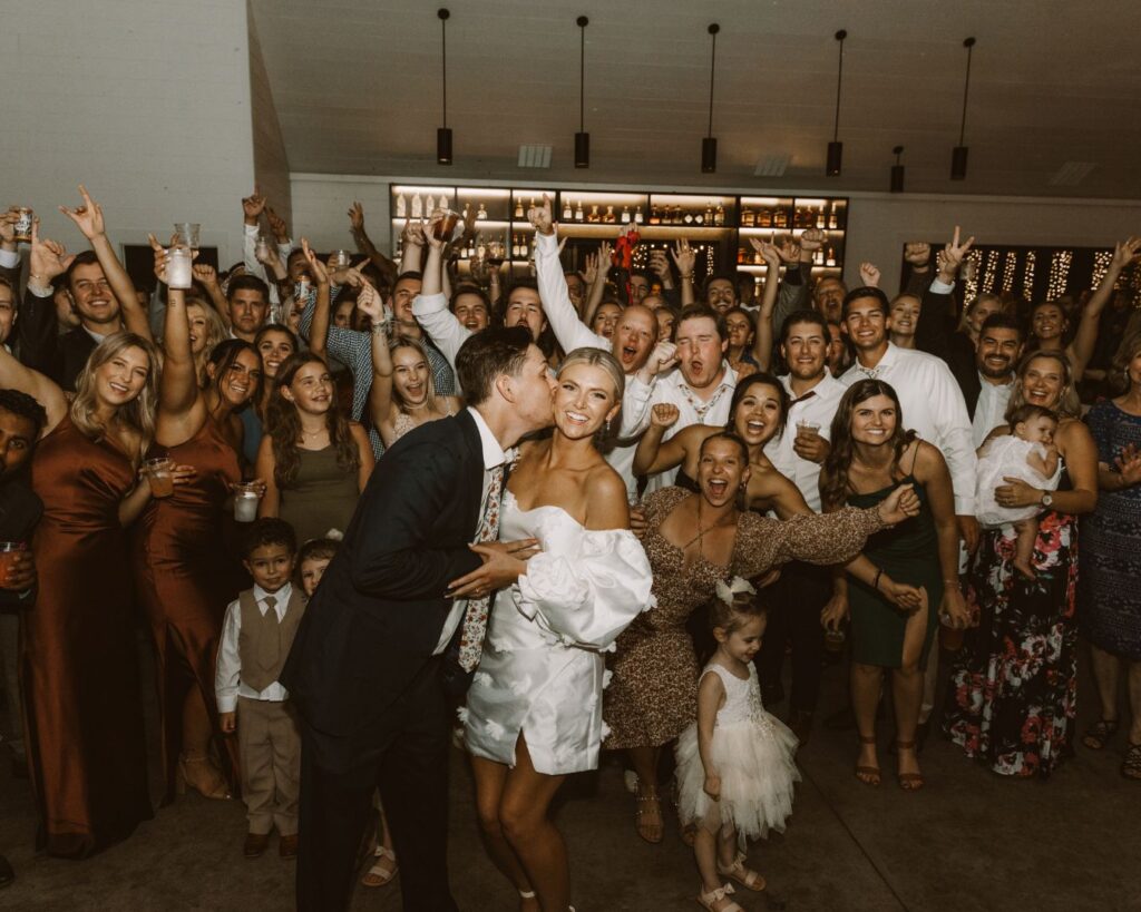 A large group photo of the bride and groom on the dance floor with a bunch of guests behind them cheering them on as he kisses her on the cheek. 