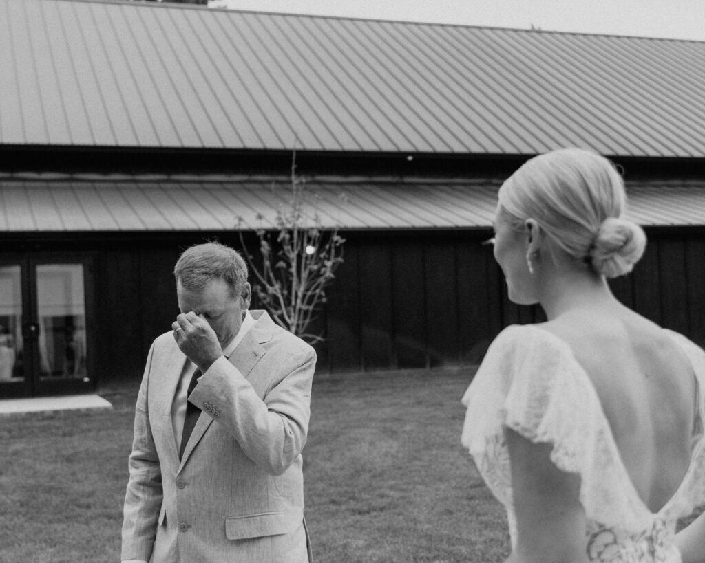 A black and white image of the father of the bride wiping his tears  when he sees his daughter for the first time in her dress on her wedding day. 