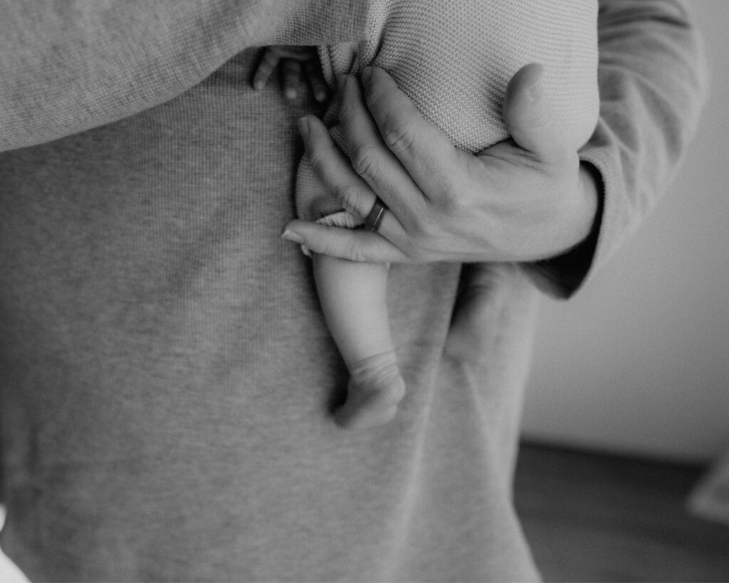 Black and white first-time father holds new born baby to his chest with his right hang on his baby's back and his ring hand under his bottom