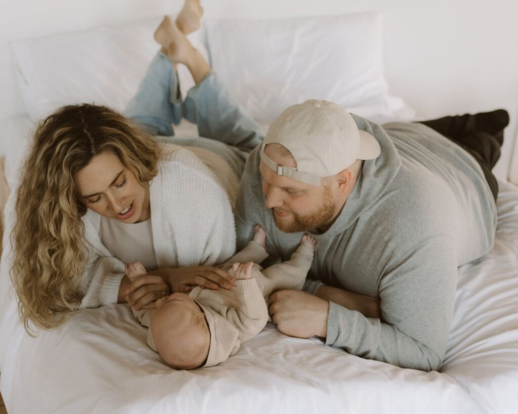 First-time mother and father lay on their bellies in bed and stare down lovingly at their baby boy who is on his back in between them