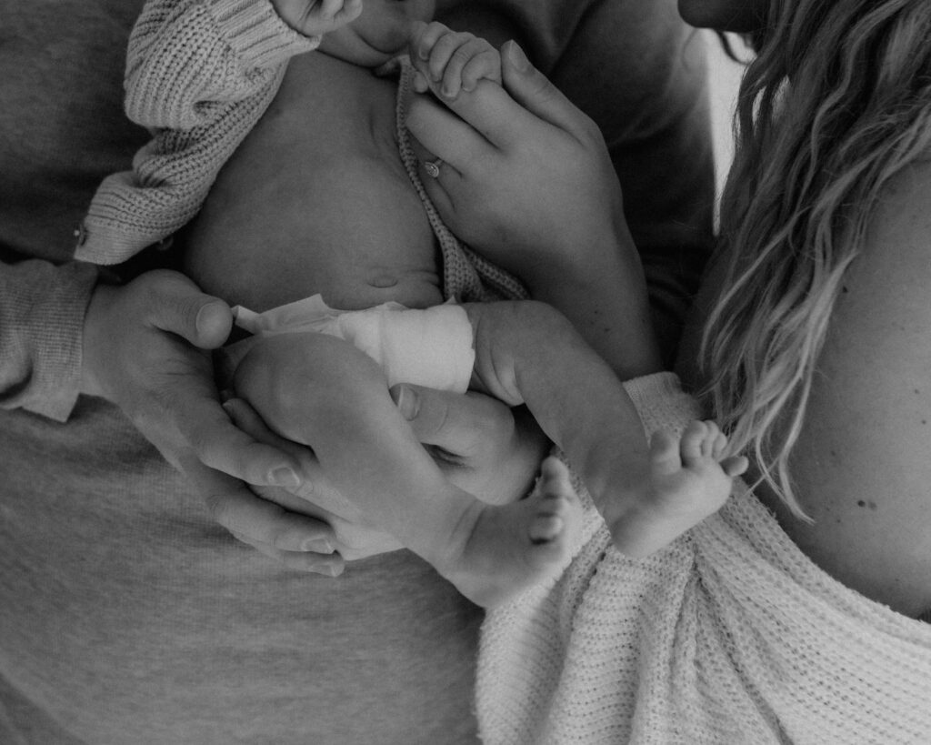 Black and white photograph of mom and dad hold their baby together while looking lovingly at him as he holds their fingers.