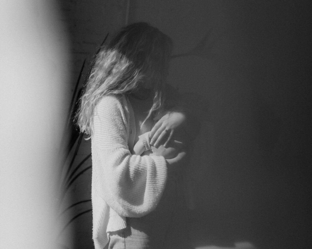 Black and white photograph of first time mother stands cradling her baby in morning light.